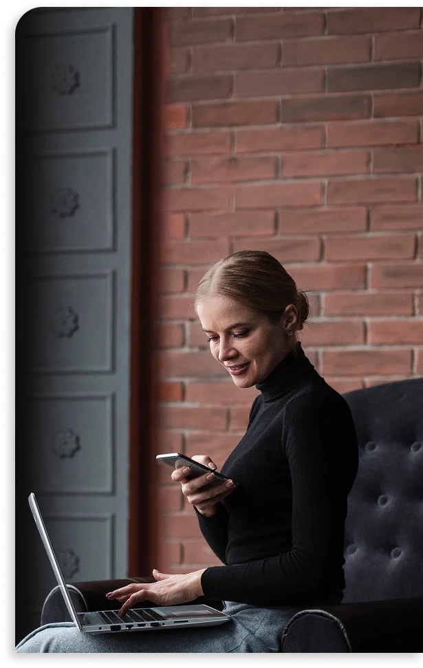 Kobieta w czarnym golfie, siedząca na ciemnym krześle, uśmiecha się, korzystając ze smartfona i laptopa. Ceglane tło nadaje wnętrzu przytulny charakter.